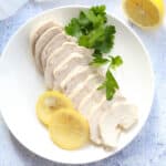 slices of poached chicken on a white plate with lemon slice and fresh parsley leaves