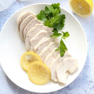 slices of poached chicken on a white plate with lemon slice and fresh parsley leaves
