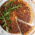 A golden-brown cauliflower frittata tortilla topped with fresh arugula rests elegantly on parchment paper. A slice has been cut and slightly pulled away, revealing its savory layers beneath the crisp exterior.