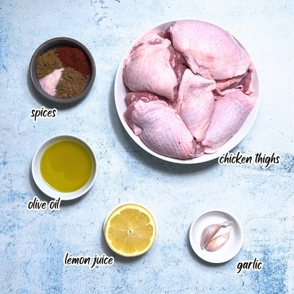 Raw chicken thighs in a bowl, surrounded by labeled bowls of spices, olive oil, lemon juice, and garlic on a light blue background—perfect ingredients for a delicious Middle Eastern Chicken Shawarma recipe.