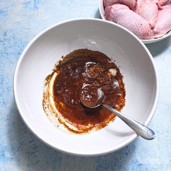 A white bowl with a metal spoon holds a reddish-brown marinade mixture, perfect for a delicious Middle Eastern Chicken Shawarma recipe. In the upper right, a small bowl contains raw chicken pieces. The background is a light blue, textured surface.