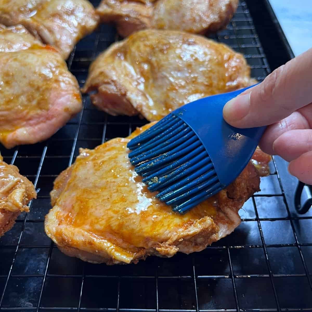 A hand uses a blue silicone brush to spread sauce on raw, seasoned chicken pieces resting on a wire rack, preparing them for a delicious Middle Eastern Chicken Shawarma recipe.
