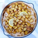 A skillet filled with roasted lemon potatoes, garnished with two lemon slices. The golden brown cubes are perfectly seasoned and coated in a light sauce.