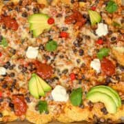 A tray of nachos supreme piled high with melted cheese, black beans, and ground meat. Topped with avocado slices, dollops of sour cream, red chili slices, and fresh cilantro, and drizzled with red salsa for an explosion of flavor.