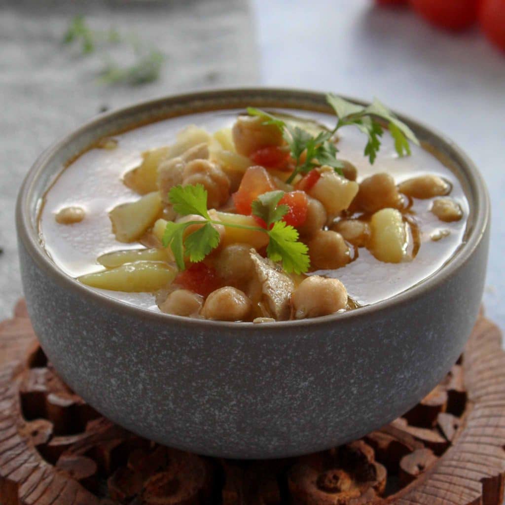 chicken soup with potatoes and chickpeas in a grey bowl on a round wooden chopping board