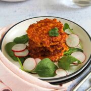 A plate of crispy fritters is stacked on fresh spinach and radish slices, garnished with a small herb. In the background, a bowl holds more fritters and a sauce. A glass of water and a fork rest nearby on a light surface.
