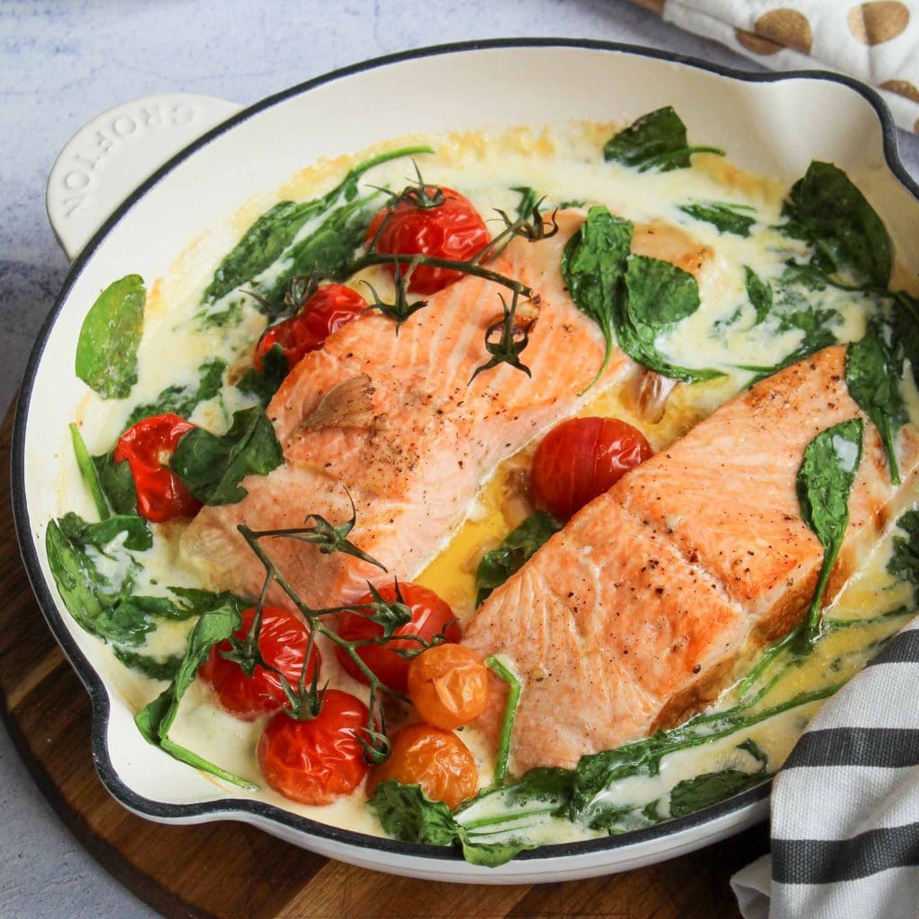 deliciously looking baked salmon with creamy sauce in a white frying pan with cherry tomatoes and spinach