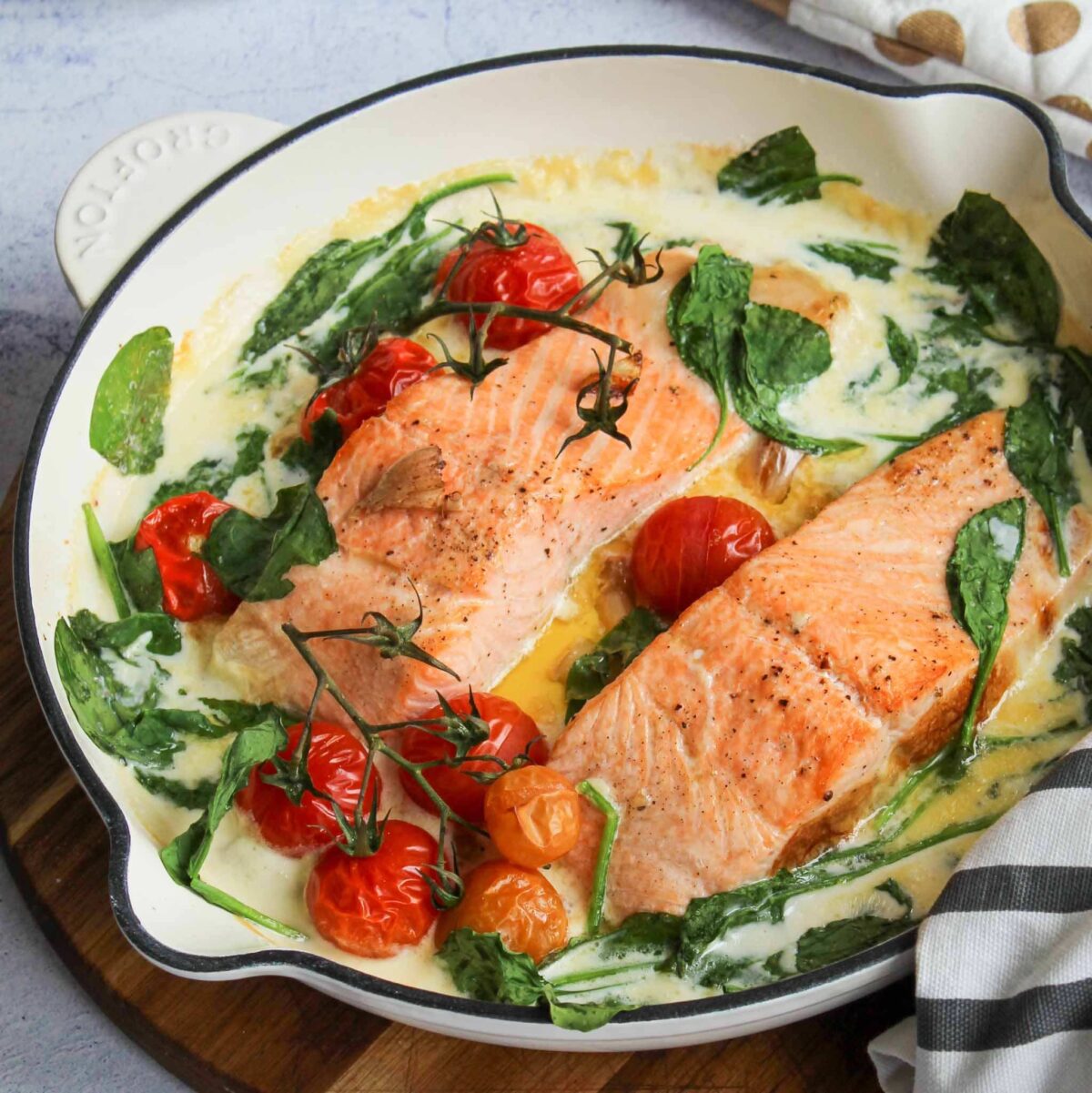 A white skillet features two cooked salmon fillets nestled among fresh spinach leaves and cherry tomatoes. Topped with a creamy white sauce, this simple and healthy, gluten-free dish rests on a wooden board with a striped cloth napkin nearby.