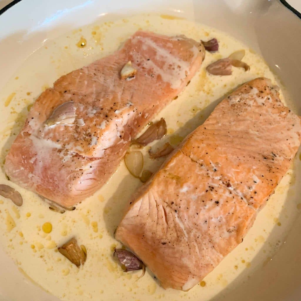 cooking step photo: 2 salmon fillets in creamy sauce and fried garlic cloves in a white frying pan