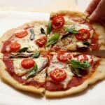 A hand holding a slice of gluten-free pizza with a thin crust, topped with tomato sauce, melted cheese, cherry tomatoes, anchovies, and fresh basil leaves. The pizza is cut into evenly-sized slices and is placed on a light surface.