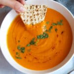 Turkish lentil soup in a white bowl, a hand is holding a slice of Turkish bread, dipping in the soup.