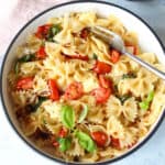 A refreshing bowl of summer pasta, featuring farfalle with cherry tomatoes, spinach, and grated cheese, garnished with fresh basil leaves. A fork rests invitingly on the right side.