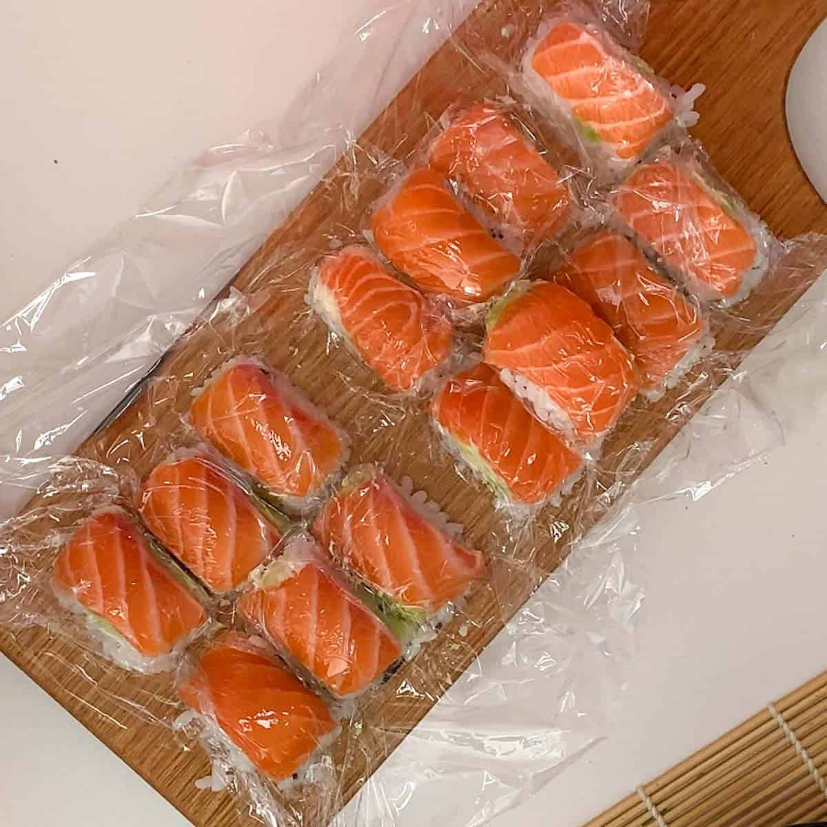 salmon nigiri covered with glad wrap on a wooden chopping board