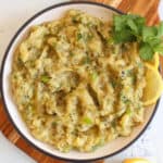 A bowl of baingan bharta garnished with cilantro and black pepper. Lemon wedges are placed on the side. The dish is on a wooden board.