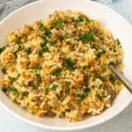 A bowl of dirty rice recipe with fried rice, mixed with minced meat, carrots, and chopped green herbs. A spoon rests in the bowl on a light-colored surface.