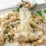 A fork lifts a portion of rice noodles mixed with minced meat, herbs, and chili flakes from a plate. The dish is garnished with fresh parsley, showcasing a flavorful combination of ingredients.