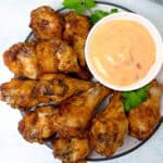 A plate of crispy dry rub baked chicken wings is served with a bowl of creamy dipping sauce. Fresh parsley garnishes the dish on a light background.