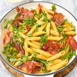 A bowl of penne pasta mixed with arugula and crispy prosciutto. The pasta is garnished with fresh greens and the prosciutto adds a crispy texture. A small bowl with grated cheese is visible in the background.