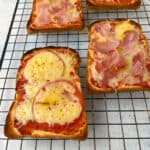 Slices of bread, transformed into delightful pizza toast, are topped with tomato sauce, melted cheese, tomato slices, and ham. They rest on a cooling rack, perfectly toasted with ingredients evenly distributed. The background features a light-colored surface.