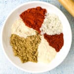A white bowl filled with neatly arranged heaps of spices, perfect for a homemade chicken taco seasoning, sits on a light blue surface. The vibrant red, green, white, and beige powders create a colorful contrast. A rolling pin is partially visible on the right edge, hinting at a kid-friendly recipe in progress.