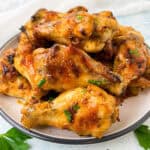 A plate of golden brown garlic butter chicken wings, glistening with glaze and garnished with chopped herbs, sits on a light-colored surface. Their crispy texture is invitingly arranged in a pile, suggesting they are freshly cooked and ready to eat.