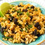 A vibrant plate of Mexican rice with black beans, garnished with cilantro and accompanied by a lime wedge. The dish is served in a teal bowl, showcasing a mix of fluffy rice, scattered beans, and fresh green herbs.