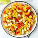 A colorful bowl of fruit salsa featuring diced mango, apple, and red bell pepper, garnished with chopped cilantro. A lime wedge is visible in the background on a light-colored surface.