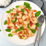 A plate of sautéed garlic shrimp garnished with red chili slices and fresh herbs. Two forks are placed beside the shrimp, and a bowl of steamed white rice is visible in the background. The dish is presented on a white tablecloth.