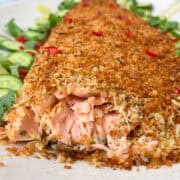 A close-up of a coconut-crusted salmon fillet with a golden breadcrumb layer, garnished with red chili flakes and served on a white plate. Fresh cucumber slices and leafy greens are visible in the background, enhancing the dish's vibrant appeal.