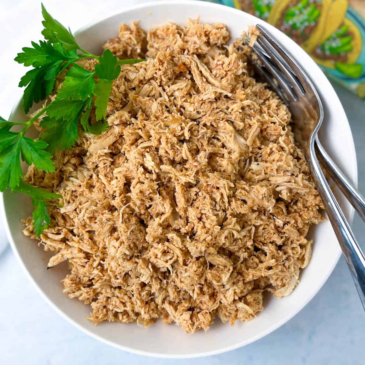 Delicious looking shredded chicken in a white bowl, served with fresh parsley and 2 forks.