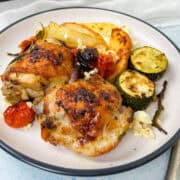 A plate of roasted chicken thighs with crispy skin, accompanied by zucchini slices, potato wedges, cherry tomatoes, and crumbled feta cheese, garnished with herbs.