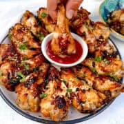 A plate of soy garlic chicken wings, garnished with sesame seeds and herbs, is arranged in a circle. A hand dips one wing into a small bowl of red sauce in the center.