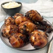 Baked chicken drumsticks with a rich glaze are piled on a plate. In the background, a bowl of rice is visible. The scene is set on a light blue surface with a white cloth nearby.