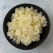A black bowl filled with cooked white butter rice sits on a light textured surface.