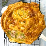 A golden-brown chicken pot pie with flaky crust, partially cut to reveal the filling of chicken and peas, sits on a black cooling rack. The crust is glossy and decorated with a leaf design, and the pie is placed on a light blue surface.