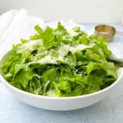 A white bowl filled with fresh green lettuce leaves, topped with grated cheese. A shiny metal cup and a white cloth are in the blurred background on a light blue surface.