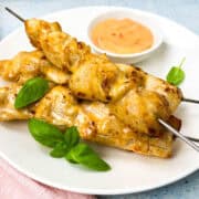 Baked bang bang chicken skewers on a white plate garnished with fresh basil leaves. A small bowl of orange dipping sauce is placed next to them. The background is a light blue surface.