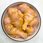 A bowl filled with raw chicken drumsticks coated in a yellow marinade, placed on a light blue surface.