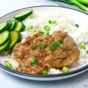 A plate of rice topped with braised adobo chicken thighs and garnished with chopped green onions. Slices of fresh cucumber are arranged on the side. The dish is served on a round, rimmed white plate.