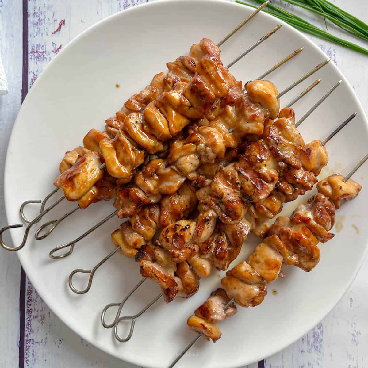 Grilled chicken skewers are arranged on a round white plate. The chicken pieces are golden brown, glazed with homemade teriyaki sauce, and threaded onto metal skewers. Green chives are visible in the background on the table.