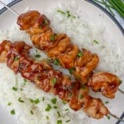 Two teriyaki glazed chicken skewers topped with chopped chives rest on a bed of white rice, served on a white plate with a black rim. Fresh chives are visible in the background.